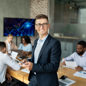 Business Leadership. Portrait Of Successful Young Businessman With Digital Tablet Posing In Office During Corporate Meeting With Collagues, Male Entrepreneur Leaning On Desk And Smiling At Camera